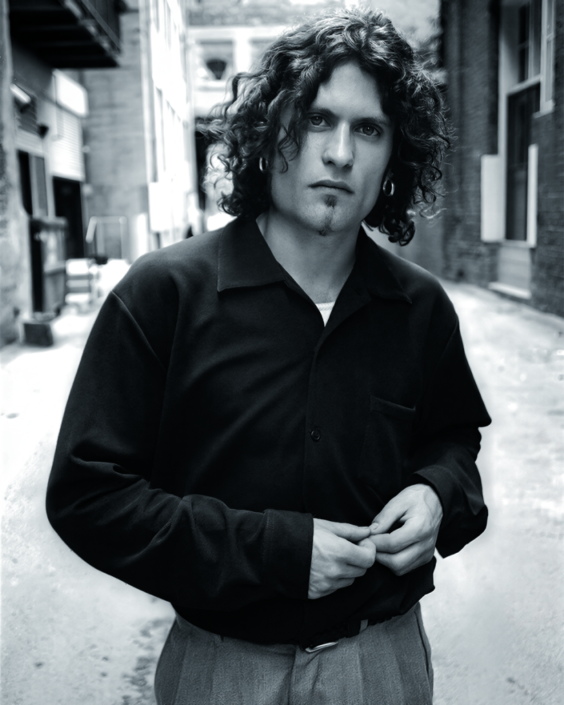 Black-and-white portrait of a curly-haired man wearing a dark shirt and hoop earring, standing in an urban alley.